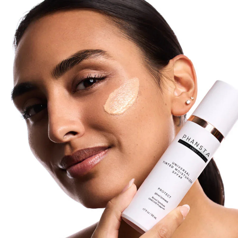Woman applying tinted moisturizer SPF 48, holding PHANSTA skincare bottle, close-up